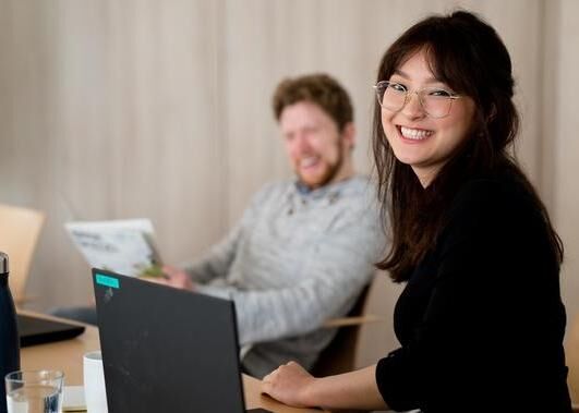 Man sieht zwei Mitarbeitende der GLS Bank vor Laptops sitzen und in die Kamera lächeln.