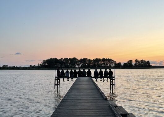 Man sieht einen Steg der geradeaus auf einen See führt. Am Ende des Stegs sieht man die Silhouette von Menschen, die dort vor dem Sonnenuntergang sitzen.